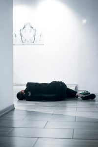 A man in casual wear lying on a tile floor in a bright, modern gallery.