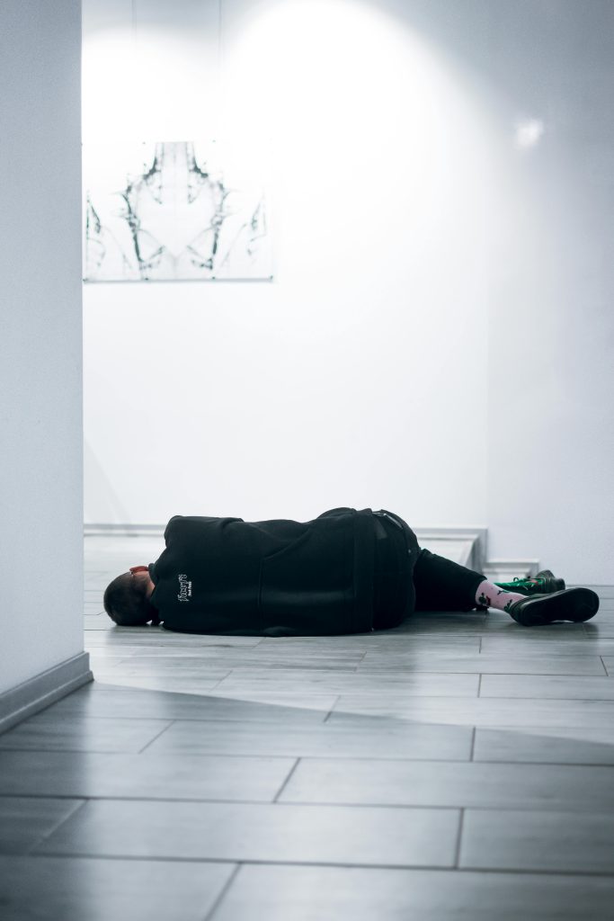 A man in casual wear lying on a tile floor in a bright, modern gallery.
